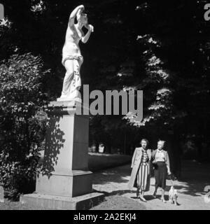 Zwei junge Frauen mit Foxterrier kommen mit einem une statue dans le parc à Stuttgart vorbei, Deutschland 1930 er Jahre. Deux jeunes femmes et un fox-terrier en vous promenant dans un jardin public à Stuttgart, Allemagne 1930. Banque D'Images