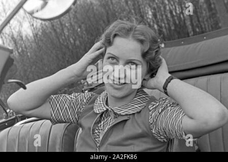 Eine junge Frau schminkt sich im Seitenspiegel votre Cabrios, Deutschland 1930er Jahre. Une jeune femme contrôler son faire dans le miroir de l'aile de son cabriolet, Allemagne 1930. Banque D'Images