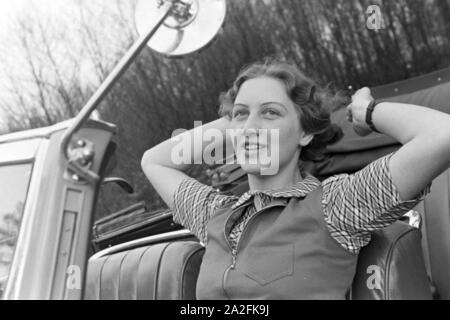 Eine junge Frau schminkt sich im Seitenspiegel votre Cabrios, Deutschland 1930er Jahre. Une jeune femme contrôler son faire dans le miroir de l'aile de son cabriolet, Allemagne 1930. Banque D'Images