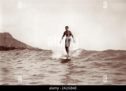 Médaille d'or olympique de natation et père de la surf, Duke Kahanamoku, une vague sur une planche en bois à Waikiki, Hawaii avec Diamond Head à l'arrière-plan. Banque D'Images