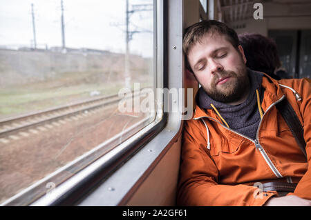 Beau jeune homme hipster dans une veste orange endormi près d'une fenêtre d'un train Banque D'Images