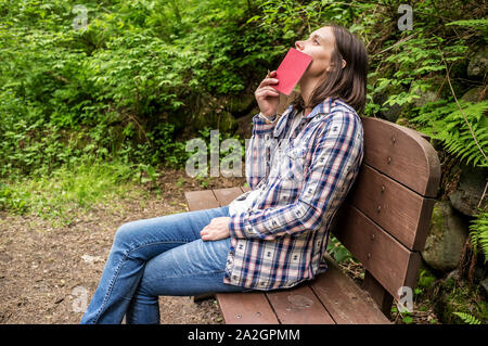 Belle femme assise sur un banc en bois dans un parc de la forêt par le chemin et a l'air rêveur le ciel, mettre un ordinateur portable à ses lèvres. Banque D'Images