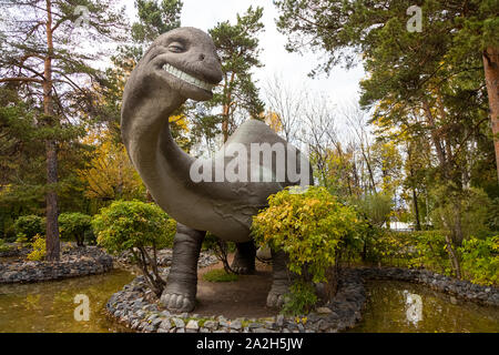Un grand brachiosaurus adultes gris regarde vers le châssis et se tient sur la pierre de la rivière, à côté de petits arbres et d'arbustes sur un automne chaud da Banque D'Images