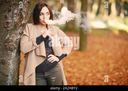 Fille dans le parc de l'automne s'appuyant sur un arbre et de fumer une cigarette électronique Banque D'Images