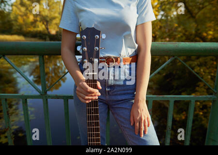 Jeune fille se tient sur le pont près de la clôture. Femme tenant une guitare acoustique par le manche de la guitare. La belle nature et rivière sur l'arrière-plan. Banque D'Images