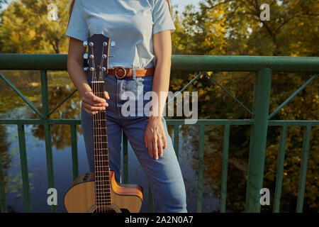 Jeune fille se tient sur le pont près de la clôture. Femme tenant une guitare acoustique par le manche de la guitare. La belle nature et rivière sur l'arrière-plan. Banque D'Images