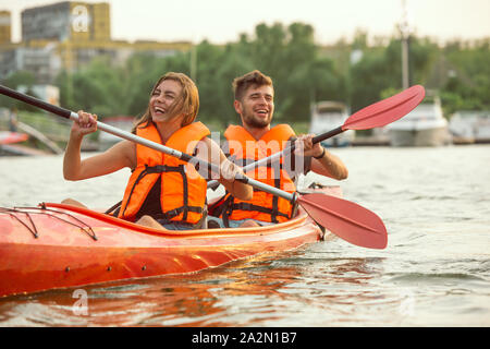 Happy young caucasian couple du kayak sur la rivière au coucher du soleil dans l'arrière-plan. Avoir du plaisir dans l'activité de loisirs. Heureux les hommes et les femmes sur le modèle laughting kayak. Le sport, les relations concept. Coloré. Banque D'Images