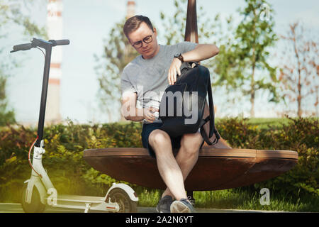 Séduisant jeune homme de parler par téléphone mobile et assis à côté d'electric scooter de coup au paysage du parc moderne. Banque D'Images