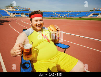 Gros homme mangeant un hamburger et de la bière dans la formation au stade. Banque D'Images