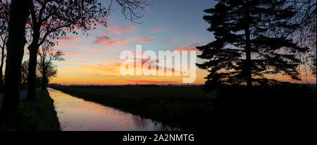 Vue panoramique d'un coucher de soleil sur le paysage hollandais. Nuage coloré reflets dans l'eau d'un canal. Silhouettes d'arbres sont visibles contre le ciel. Banque D'Images