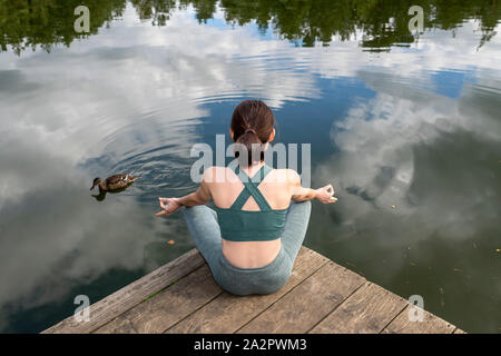 Woman practicing yoga on une jetée par un lac, vue de dos assis. Banque D'Images