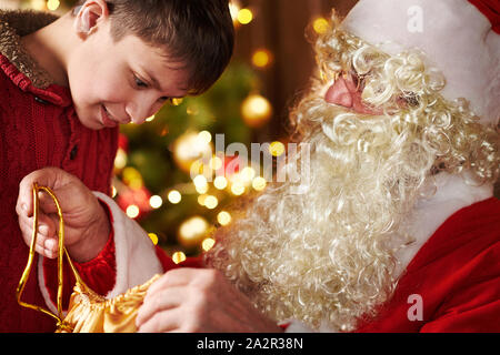 Le Père Noël ouvre un sac et donne des cadeaux aux enfants, assis près de l'arbre de Noël décoré avec des lumières - Joyeux Noël et bonnes fêtes ! Banque D'Images