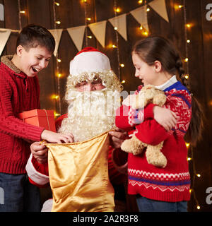 Le Père Noël ouvre un sac et donne des cadeaux aux enfants, assis près de l'arbre de Noël décoré avec des lumières - Joyeux Noël et bonnes fêtes ! Banque D'Images