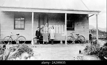 Négatif - Groupe de la famille des vélos à l'extérieur & Farm House, Mooralla, Victoria, vers 1935, une femme, un petit garçon et une fille debout devant une maison. La femme et la petite fille portent des robes, et le petit garçon est portant une chemise avec un short. Il y a deux vélos, un pour une fille, appuyé contre la maison. La maison a deux grandes fenêtres de chaque côté de la porte, et les rideaux peuvent être vus à travers la fenêtre sur la gauche. La maison est en bois de style victorien avec un toit de fer Banque D'Images