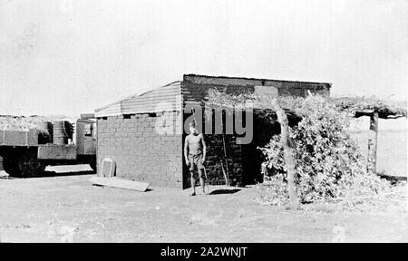 - Négatif, Tanami Territoire du Nord, 1937, un jeune homme debout à l'entrée d'un bâtiment en briques de boue. Il y a une véranda de broussailles appuyé par les troncs des arbres. Il y a un camion transportant des fûts d'huile à l'arrière-plan Banque D'Images