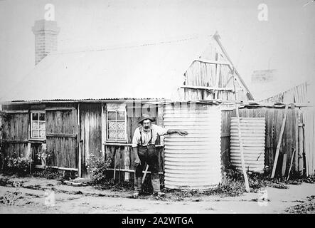 Négatif - Bairnsdale District, Victoria, vers 1890, un homme âgé à l'extérieur d'une cabane d'écorce qui a un toit en tôle ondulée. Il est debout avec sa main sur l'un des deux réservoirs d'eau Banque D'Images