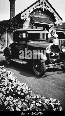 Négatif - Geelong, Victoria, vers 1930, une femme portant un manteau garni de fourrure debout à côté d'un Ford un modèle coupé. Il y a une maison en bois dans l'arrière-plan Banque D'Images