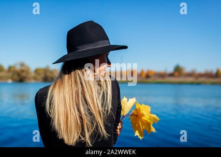 Automne vibes. Young woman relaxing by lake holding jaune feuilles d'érable Banque D'Images