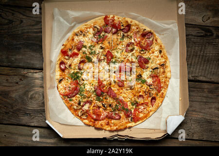 Ronde au four pizza avec saucisses fumées, champignons, tomates, fromage et de l'aneth dans une boîte en carton sur une table en bois, de l'alimentation est coupée en portions, t Banque D'Images