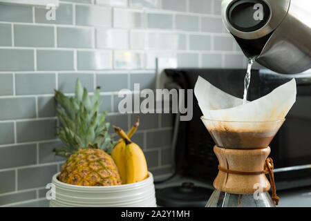 Gros plan d'une bouilloire électrique versant de l'eau chaude dans un récipient à café avec un bol à fruits en arrière-plan. Banque D'Images