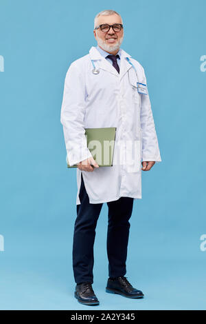 Portrait of mature médecin en blouse blanche et à lunettes holding folder and smiling at camera sur fond bleu Banque D'Images