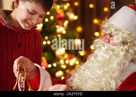 Le Père Noël ouvre un sac et donne des cadeaux aux enfants, assis près de l'arbre de Noël décoré avec des lumières - Joyeux Noël et bonnes fêtes ! Banque D'Images