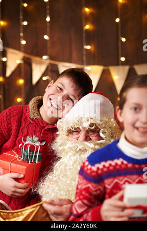 Le Père Noël ouvre un sac et donne des cadeaux aux enfants, assis près de l'arbre de Noël décoré avec des lumières - Joyeux Noël et bonnes fêtes ! Banque D'Images