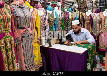 Un tailleur dans son atelier assis en face à une machine à coudre avec des robes colorées à Muscat, Oman Banque D'Images