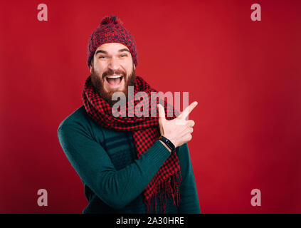 Heureux homme barbu, aisde de pointage montrant quelque chose sur fond rouge. Vacances d'hiver. Banque D'Images