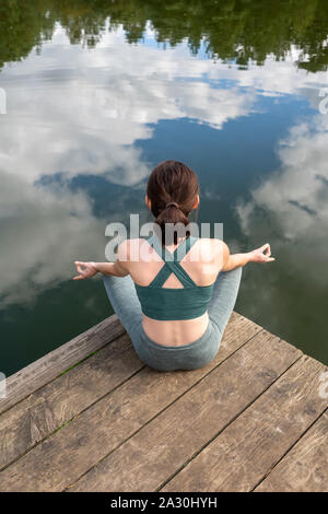 Woman practicing yoga on une jetée par un lac, vue de dos assis. Banque D'Images