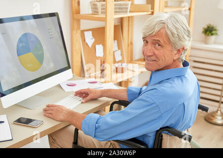 Portrait of senior man sitting on fauteuil roulant et à la caméra à tout en travaillant avec des graphiques sur ordinateur à la table Banque D'Images