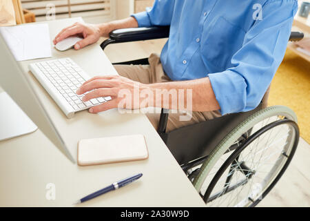 Close-up of senior mobilité homme assis en fauteuil roulant à la table et de la saisie sur clavier d'ordinateur tout en travaillant au bureau Banque D'Images