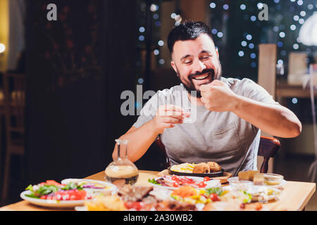 Homme boit de la vodka sur une grande table avec des aliments gras et des collations Banque D'Images