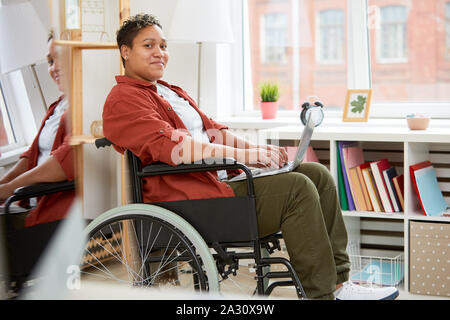 Portrait de jeune femme africaine handicapées assis dans fauteuil roulant et looking at camera while using laptop computer at home Banque D'Images