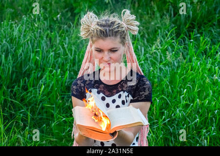 La jeune fille est titulaire d'un livre brûlant dans ses mains. Une jeune femme dans une forêt brûle un livre. Banque D'Images