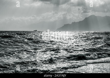 Paysage marin noir et blanc avec un seul navire et les montagnes d'ossature sur beau ciel nuages de fond avec les rayons du soleil Banque D'Images