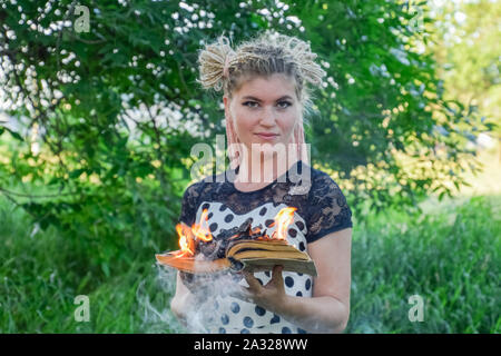 La jeune fille est titulaire d'un livre brûlant dans ses mains. Une jeune femme dans une forêt brûle un livre. Banque D'Images