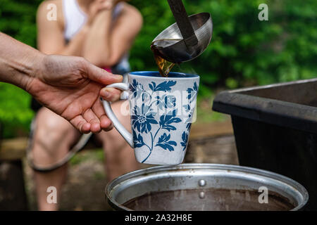 Une vue en gros plan sur les mains d'une personne au service de thé naturel et sain une casserole sur un feu de camp au centre d'une forêt sacrée. Banque D'Images