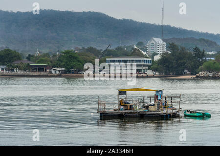 Si Racha, Thaïlande - Mars 16, 2019 : Gros plan du radeau de pêche flottant sur l'eau de la baie de la Thaïlande avec la ville et certains immeubles blancs et boisées de hil Banque D'Images