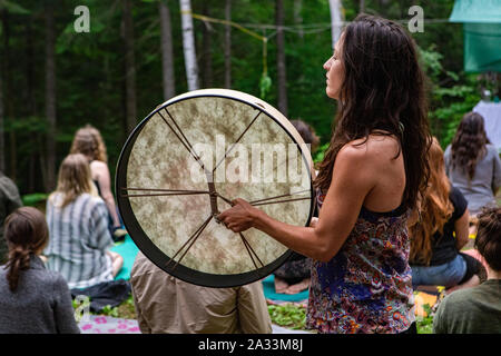 Un gros plan et profil latéral d'une femme de race blanche de la lecture d'un grand tambour traditionnels autochtones comme un groupe mixte de personnes méditer dans les bois. Banque D'Images