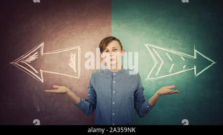 Perplexe teenage boy propagation les bras tendus, l'équilibre entre les mains le geste, deux options au choix à droite ou à gauche, la prise de concept. Un tableau noir et de Split Banque D'Images