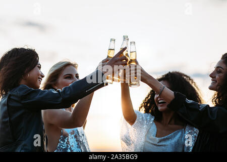 Multi-ethnic friends toasting bières au coucher du soleil. Les femmes célébrant avec bières dans la soirée. Banque D'Images