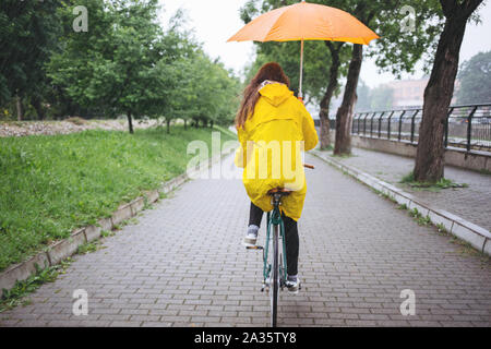 Balade à vélo sous la pluie. Femme en imperméable riding bicycle et holding umbrella Banque D'Images