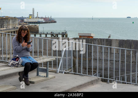 Une femme d'âge moyen assis sur un banc, à un point de vue de la mer à la recherche de son téléphone portable ou un téléphone cellulaire Banque D'Images