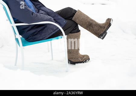 L'homme dans des vêtements chauds et des bottes de feutre, s'assoit et se détend dans une chaise qui se trouve dans la neige à l'extérieur, l'hiver glacial à la campagne. Close-up. Banque D'Images