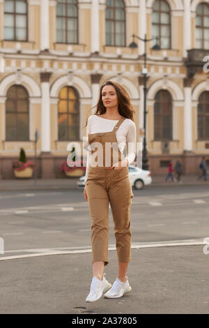 Femme élégante dans brown classement global, poser et marcher à la rue de la ville au coucher du soleil. Belle fille avec de longs cheveux bruns ondulés Banque D'Images