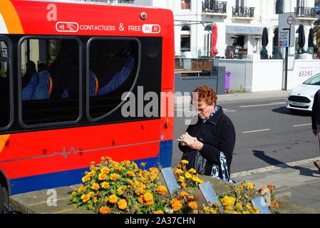 Une dame âgée seule debout à côté d'un bus à l'aide d'un smart phone, sur le front de Worthing Sussex England UK Banque D'Images