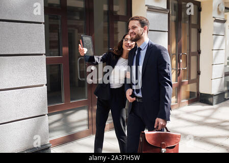 Attractive brunette woman holding a tablet lunettes en montrant à un homme barbu beau cartable en cuir marron avec la photo en gros plan. Banque D'Images