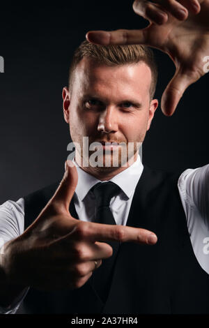 Les jeunes bel homme en chemise blanche, gilet noir qui cadre avec ses mains sur fond noir, la photo en gros plan sur fond noir isolé. Banque D'Images