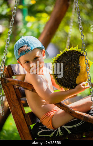 Little Boy holding tête de tournesol mûres. Mignon petit garçon dans une casquette de baseball est assis sur une balançoire Banque D'Images